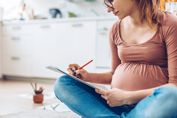 There are some alert signs during the first trimester you need to have in mind. A pregnant woman taking notes about her first trimester while sitting on the floor.