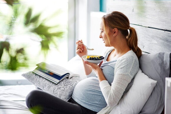Try to opt for healthy food for your pregnancy cravings. A woman eating fruit on the couch while reading.